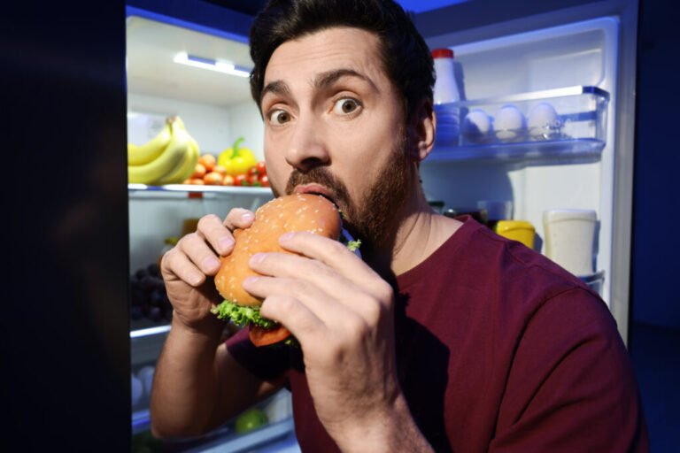 Man eating burger near refrigerator in kitchen at night Bad habit